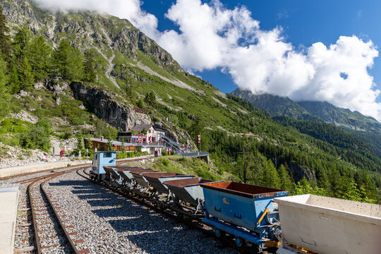 View of the VerticAlp Emosson mountain railway with vintage mining wagons on tracks, set against lush alpine slopes, rocky cliffs, and blue sky in Le Chatelard, Fribourg, Switzerland.