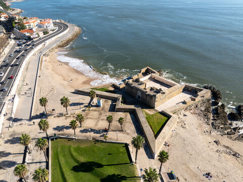 Aerial view of Forte de Sao Bruno, a historic star-shaped fortress situated on the coastline with sandy beaches, palm trees, and a coastal road in Caxias, Lisbon, Portugal.