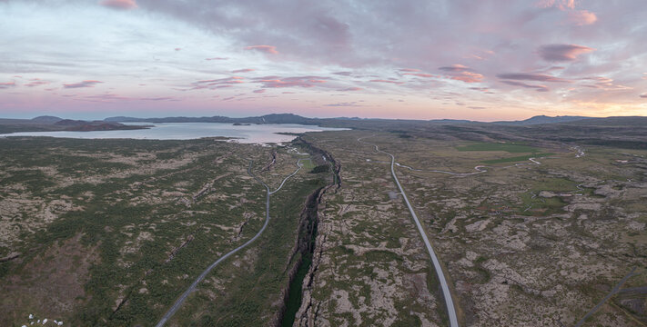 Aerial view of the tectonic rift valley at Thingvellir National Park with mossy lava fields, a winding road, and a distant lake under a sunset sky in Selfoss, Blaskogabyggd, Iceland.