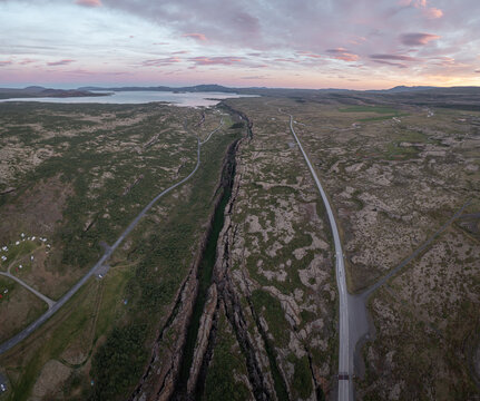 Aerial view of the Almannagja tectonic rift in Thingvellir National Park, featuring lava fields, mossy terrain, and a road under a soft sunset sky in Selfoss, Blaskogabyggd, Iceland.