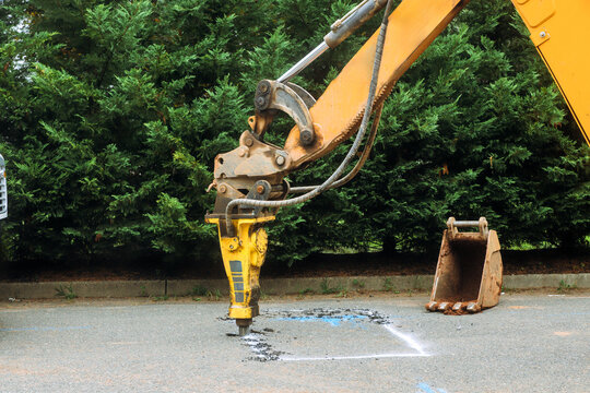 Yellow excavator digs into asphalt on road uses jackhammer while trees line daytime.