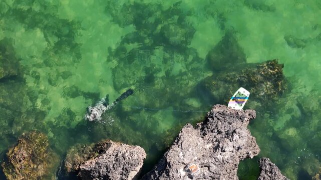 Aerial view of a scuba diver swimming in clear green water near rocky cliffs with a small board on the shore in Cascais, Lisbon, Portugal.