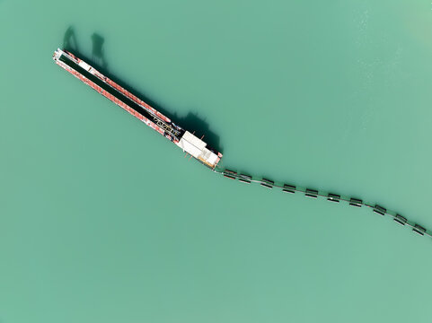 Aerial view of a long industrial barge with a floating containment boom trailing behind on the turquoise water of the Mondego River in Coimbra, Coimbra District, Portugal.