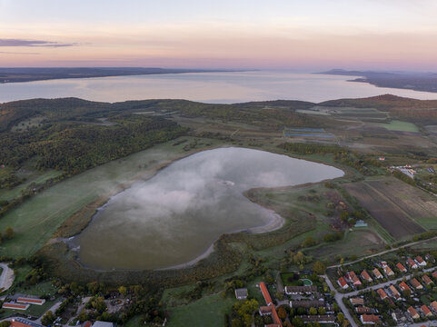 Aerial view of the Inner Lake in Tihany, surrounded by rolling hills, green fields, and residential houses, with Lake Balaton in the background under a soft sunrise sky. Tihany, Hungary.