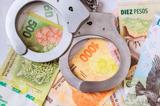 Handcuffs lie atop Argentine pesos, with various banknotes on suggesting crime connection.