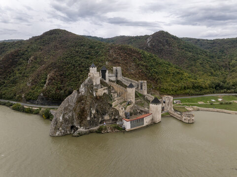 Aerial view of Golubac Fortress perched on a rocky cliff overlooking the Danube River, surrounded by lush green hills under a cloudy sky, Golubac, Branicevski Okrug, Serbia.