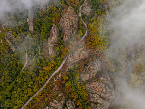 Aerial view of Belogradchik Rocks surrounded by autumn forest and a winding road partially obscured by mist in Belogradchik, Vidin, Bulgaria.