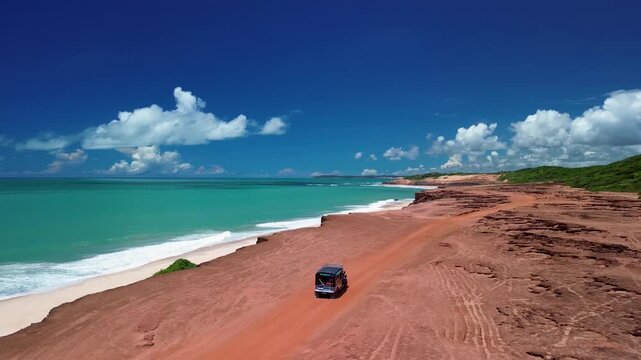 Aerial view of a vehicle driving along the red dirt road atop the cliffs overlooking the turquoise ocean at Pipa Beach, Rio Grande do Norte, Brazil.