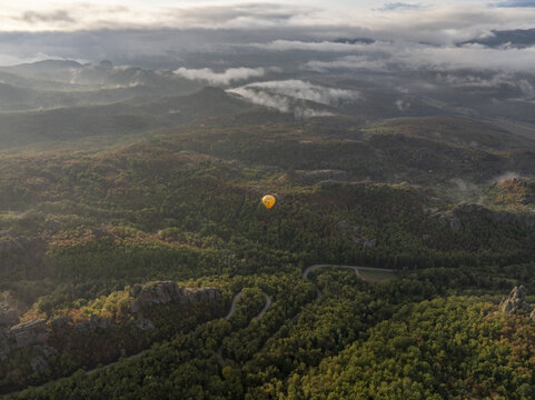 Aerial view of a yellow hot air balloon floating above the unique rock formations and lush forests of the Belogradchik Rocks at sunrise in Belogradchik, Vidin, Bulgaria.