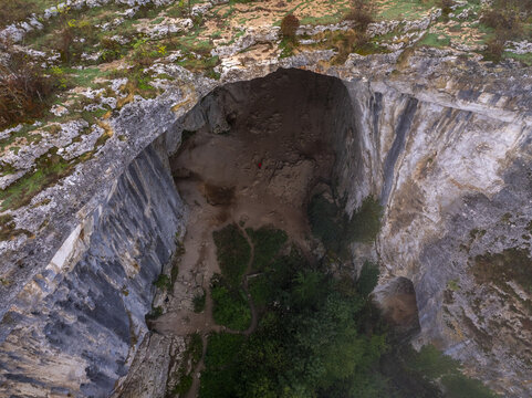 Aerial view of Prohodna Cave with its massive limestone arch, rocky cliffs, lush greenery, and natural light filtering through the cave openings in Karlukovo, Lovech, Bulgaria.