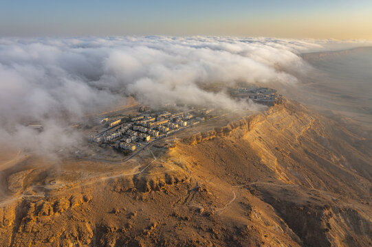 Aerial view of Mitzpe Ramon crater partially covered by low-hanging clouds at golden hour, showcasing the desert landscape and town in Mitzpe Ramon, South District, Israel.
