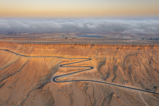 Aerial view of the winding road through the desert landscape of Makhtesh Ramon at sunset with low clouds over the crater rim in Mitzpe Ramon, South District, Israel.