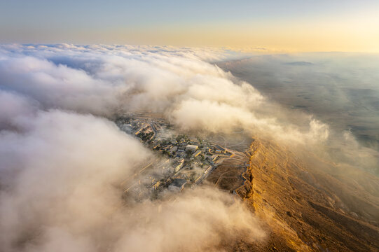 Aerial view of Mitzpe Ramon town perched on the edge of the Makhtesh Ramon crater, partially obscured by thick clouds during the golden hour in Mitzpe Ramon, South District, Israel.