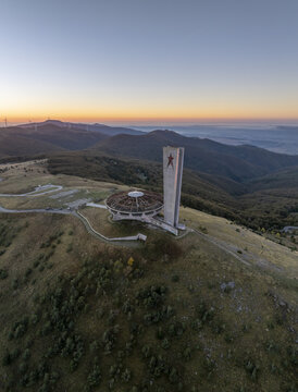 Aerial view of the Buzludzha Monument, a brutalist structure with a circular roof and tall tower featuring a red star, set against rolling hills in Stara Zagora, Bulgaria.