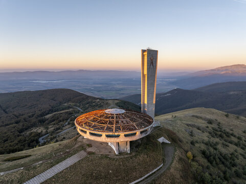 Aerial view of the iconic Buzludzha Monument, a brutalist structure perched on a mountain peak at sunset, surrounded by rolling hills and autumn forests in Stara Zagora, Bulgaria.