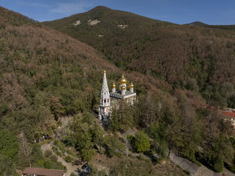 Aerial view of Shipka Memorial Church nestled in a dense forest with golden domes and a tall bell tower against a mountain backdrop in Shipka, Stara Zagora, Bulgaria.