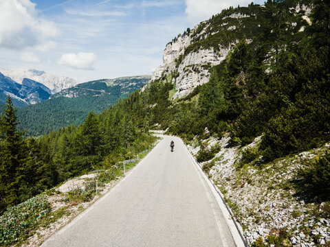 Aerial view of a cyclist riding on a mountain road through the Dolomites surrounded by pine forests and rocky peaks in Italy.