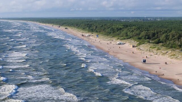 Aerial view of the sandy Palanga beach with rolling Baltic Sea waves, lush pine forest, and people enjoying the summer day in Palanga, Klaipeda County, Lithuania.