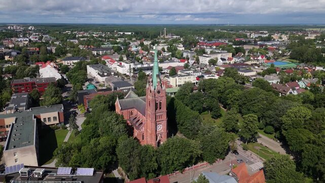 Aerial view of the red brick Church of the Assumption of the Virgin Mary surrounded by lush green trees and urban rooftops in Palanga, Klaipeda County, Lithuania.