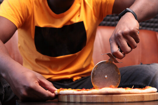 Close-up of hands of man with dark skin tone, slicing fresh pizza with metal wheel on wooden board against background of leather sofa