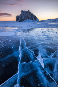 View of the frozen surface of Lake Baikal with deep ice cracks and a rocky island in the distance under a soft sunset sky in Irkutsk, Irkutsk Oblast, Russia.