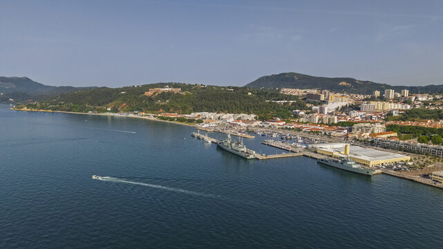 Aerial view of the harbor and coastline with naval ships docked at the pier, surrounded by urban buildings and hills in the background in Setubal, Setubal, Portugal.
