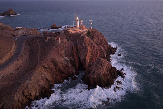 Aerial view of Lighthouse de Cabo de Gata perched on rocky cliffs above the sea at dusk, with waves crashing against the shore and a winding coastal road in San Jose, Andalucia, Spain.