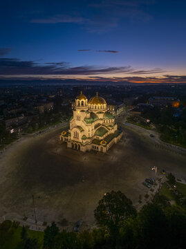Aerial view of the illuminated Patriarchal Cathedral St. Alexander Nevsky at dusk, surrounded by the city square and urban landscape in Sofia, Sofia City Province, Bulgaria.