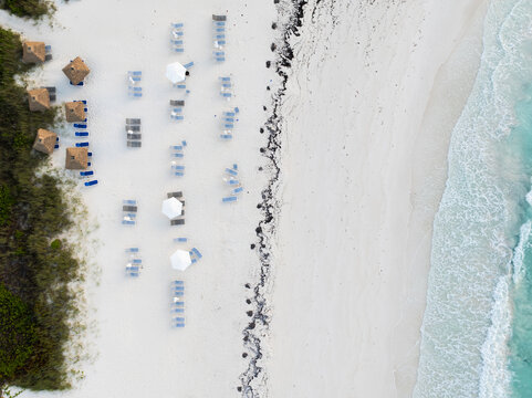 Aerial view of a pristine white sand beach with rows of lounge chairs and umbrellas, bordered by lush green vegetation and turquoise ocean waters in Exuma, Bahamas.