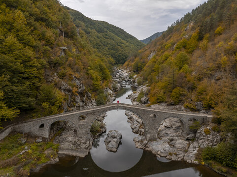 Aerial view of Devil's Bridge spanning the Arda River in a lush valley with autumn foliage and rocky terrain in Kardzhali Province, Bulgaria.
