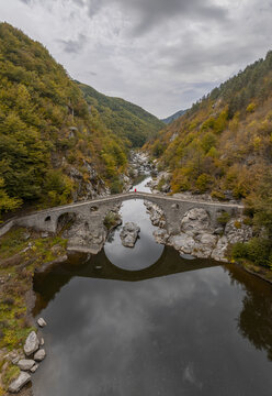 Aerial view of the historic stone Devil's Bridge arching over the Arda River, surrounded by autumn-colored forested hills and rocky banks in Kardzhali Province, Bulgaria.