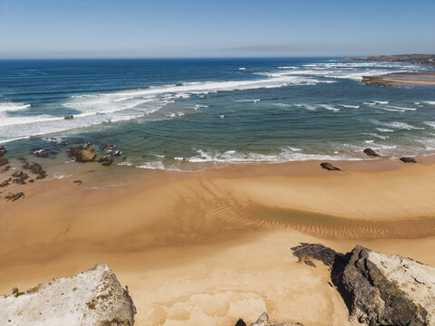 Aerial view of the sandy beach and rocky coastline of Vila Nova de Milfontes with waves crashing on the shore of the Atlantic Ocean in Vila Nova de Milfontes, Beja District, Portugal.