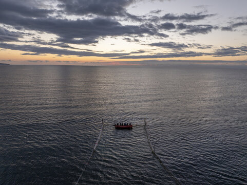 Aerial view of a small red boat with people on board floating in the calm sea at sunset near the historic town of Nessebar, with a dramatic cloudy sky in Nessebar, Burgas, Bulgaria.