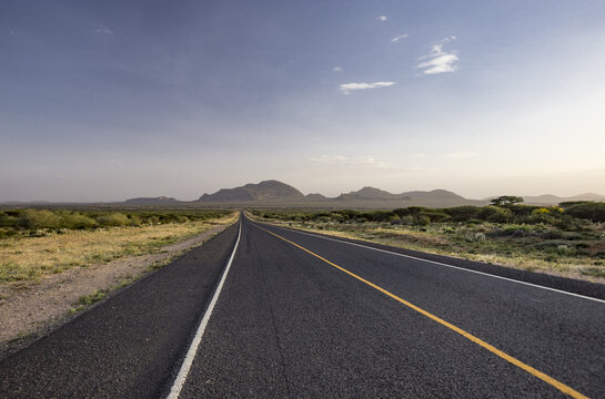 View of a long asphalt road stretching towards the Oldoinyo Sabachi mountain through the dry savanna landscape of Samburu County, Kenya.