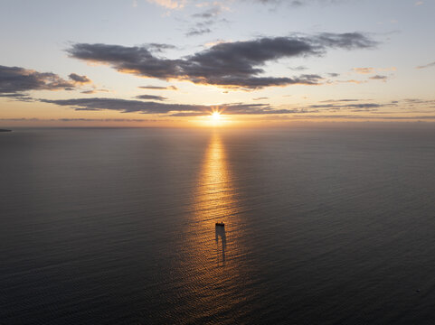 Aerial view of a small boat sailing on the calm sea during a golden sunset with a vibrant sky and sun reflection on the water near Nessebar, Burgas, Bulgaria.