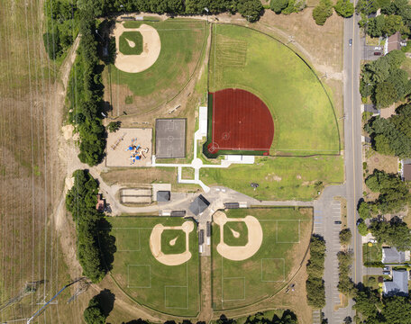 Aerial view of a sports complex featuring multiple baseball diamonds with red clay infields, a basketball court, and a playground in Commack, New York, United States.