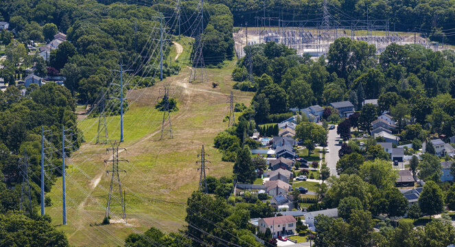Aerial view of high-voltage power transmission lines and a substation situated within a suburban neighborhood with lush trees and residential houses in Commack, New York, United States.