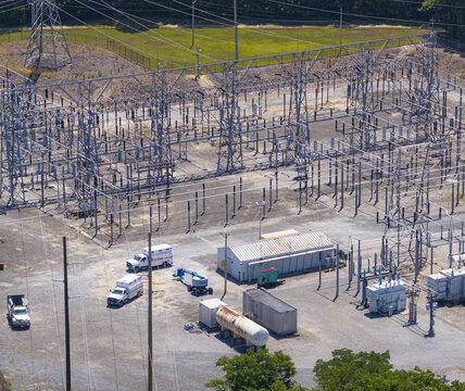 Aerial view of an electrical substation with steel lattice structures, power lines, and utility trucks parked on a gravel lot in Commack, New York, United States.