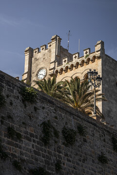 View of the historic stone clock tower of the Cathedral of Ciutadella rising above a weathered stone wall with palm trees under a clear blue sky in Ciutadella, Balearic Islands, Spain.