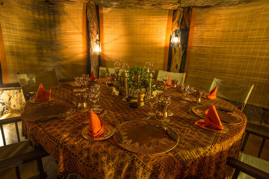 View of a dining table set for dinner in a rustic safari lodge with warm lantern lighting, patterned tablecloth, and orange napkins in Laikipia County, Kenya.