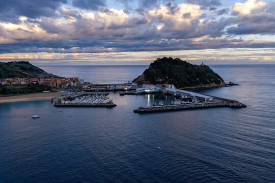 Aerial view of Getariako portua with fishing boats docked in the harbor, surrounded by the coastline and the San Anton hill under a cloudy sky in Getaria, Basque Country, Spain.