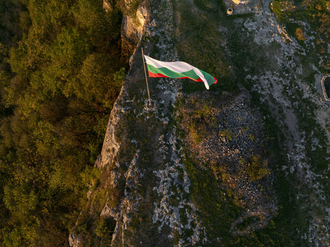 Aerial view of the Bulgarian flag waving on a pole atop the rocky cliffs of Ovech Fortress, surrounded by lush forest and rugged terrain at sunset in Provadia, Varna, Bulgaria.