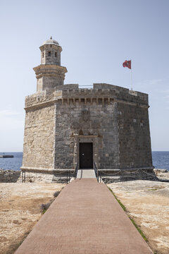 View of the historic Castell de Sant Nicolau stone tower with its entrance walkway and a red flag flying against a clear blue sky in Ciutadella, Balearic Islands, Spain.