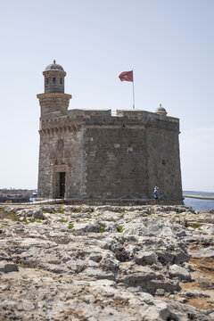 View of the historic stone watchtower Castell de Sant Nicolau with a flag flying on top, set against a clear blue sky and rocky coastline in Ciutadella, Balearic Islands, Spain.