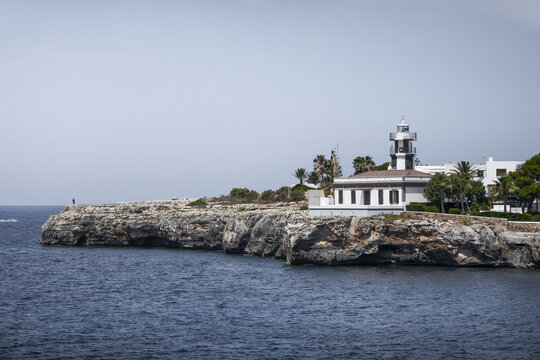 View of the Far de sa Farola lighthouse perched on the rocky coastline with palm trees and clear blue sky in Ciutadella, Balearic Islands, Spain.