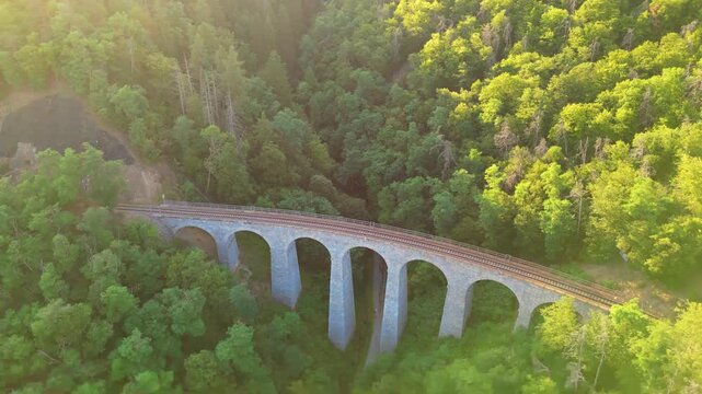 Aerial view of the historic Zampach viaduct railway bridge spanning a lush green valley during golden hour in Jilove u Prahy, Stredocesky kraj, Czechia.