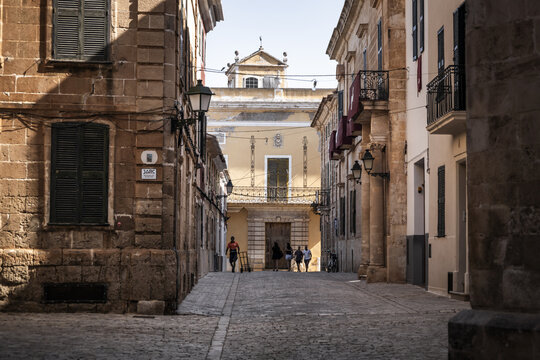 Ciutadella, Balearic Islands, Spain - 26 June 2026: View of the historic streets of Ciutadella with traditional stone architecture, warm sunlight, and people walking along the cobblestone path.