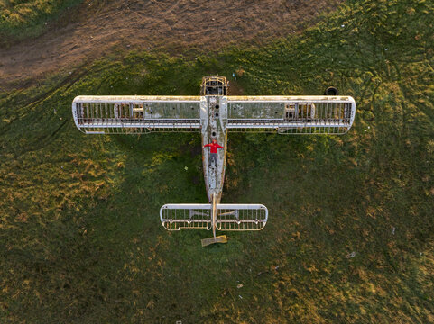 Aerial view of an abandoned airplane with a person in a red jacket standing on the fuselage in a grassy field during golden hour in Bulgaria.