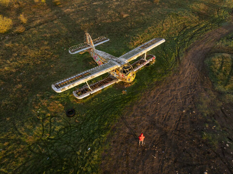 Aerial view of an abandoned biplane with graffiti on its fuselage resting in a grassy field with tire tracks and a person in a red jacket standing nearby in Bulgaria.