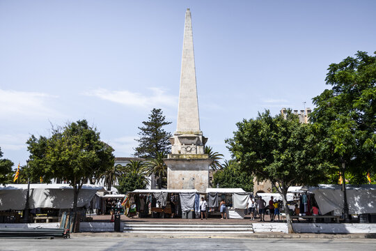 Ciutadella, Balearic Islands, Spain - 26 June 2026: View of the obelisk in Placa des Born surrounded by market stalls, trees, and people under a clear blue sky.